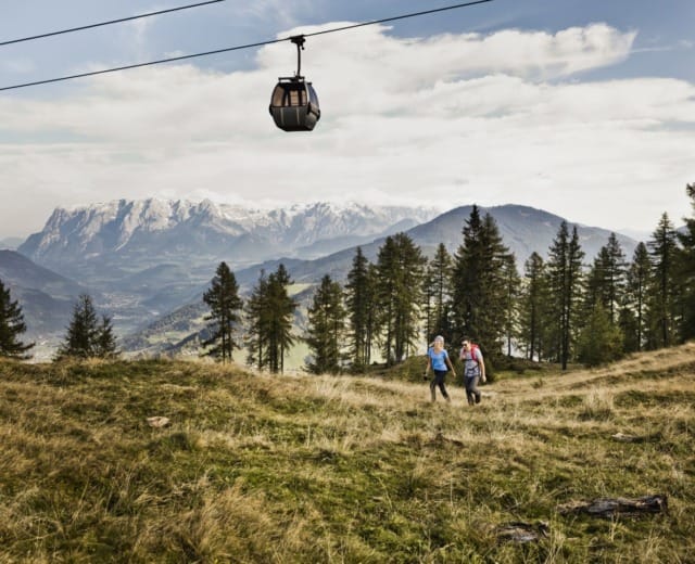 Dva turisté se procházejí po travnaté alpské louce, zatímco jim nad hlavou kloužou Letní lanovky sv. Jana v Salcbursku.