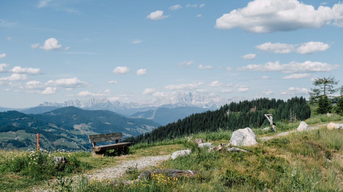Dřevěná lavička na travnatém kopci v St. Johann v Salcbursku s výhledem na vzdálené hory a oblohu.