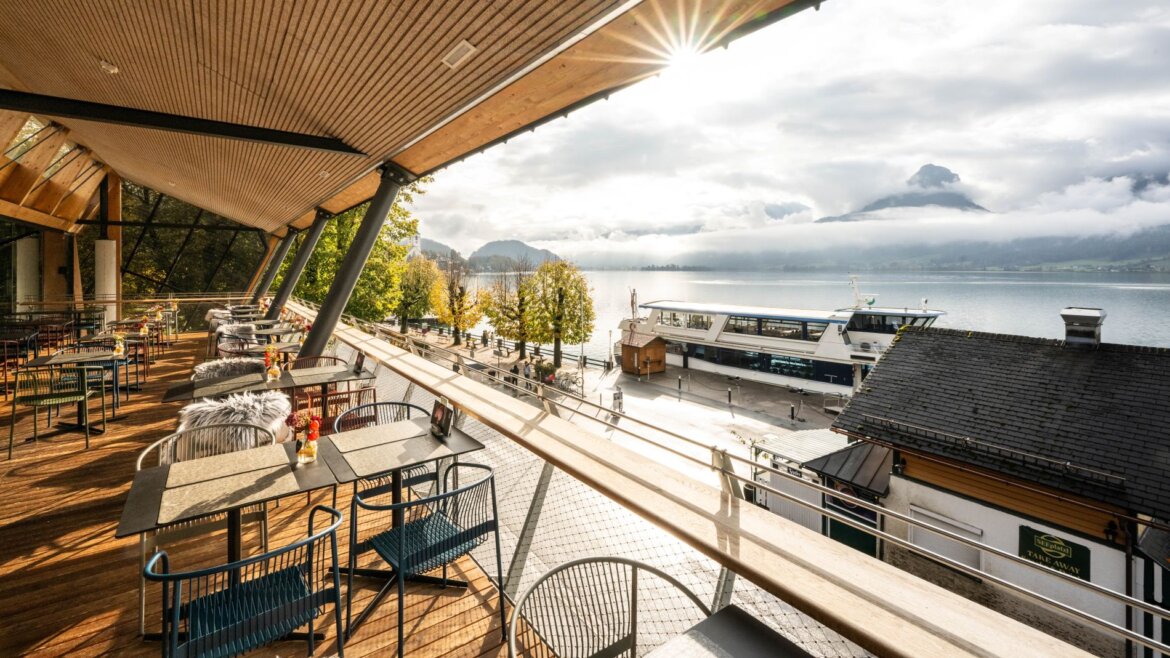Sluncem zalitá terasa restaurace ErlebnisQuartier u jezera s výhledem na hory Salzkammergutu, molo a podzimní scenérii.