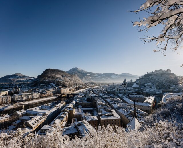 Salzburg_City_Winter_062 Zasněžená městská krajina s horami, jasnou modrou oblohou a pohledem na Hohensalzburg