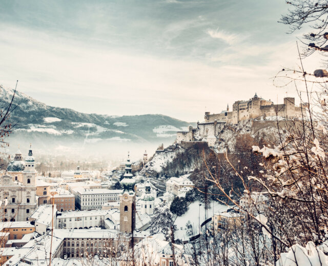 Salcburk se zasněženými střechami kostelů a domů s dominantou, pevností Hohensalzburg