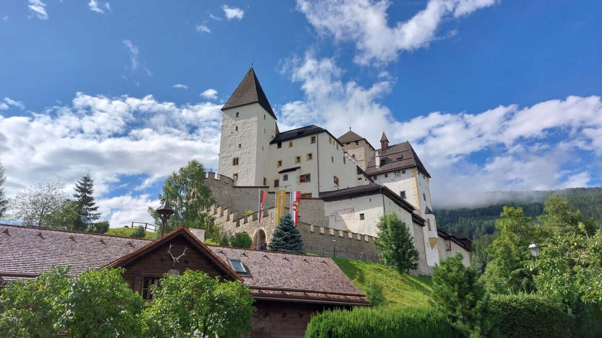 Středověký hrad Hrad Mauterndorf s věžemi na kopci, modrá obloha, mraky, stromy a dřevěný dům před ním. (Zvětšený pohled)