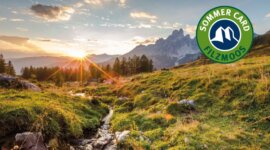 filzmoos-sommercard Východ slunce nad svěží horskou krajinou s potokem; v rohu logo Letní karty Filzmoos.