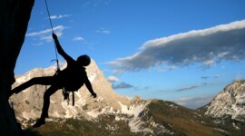 Silueta horolezce lezoucího po skalní stěně v Lezecké oblasti Filzmoos, s horami a modrou oblohou za zády.