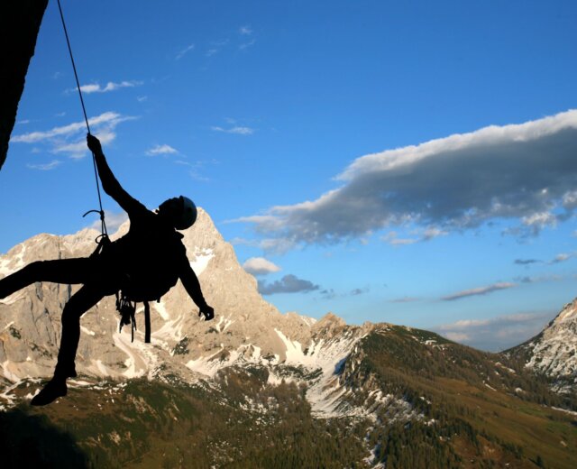 Silueta horolezce lezoucího po skalní stěně v Lezecké oblasti Filzmoos, s horami a modrou oblohou za zády.