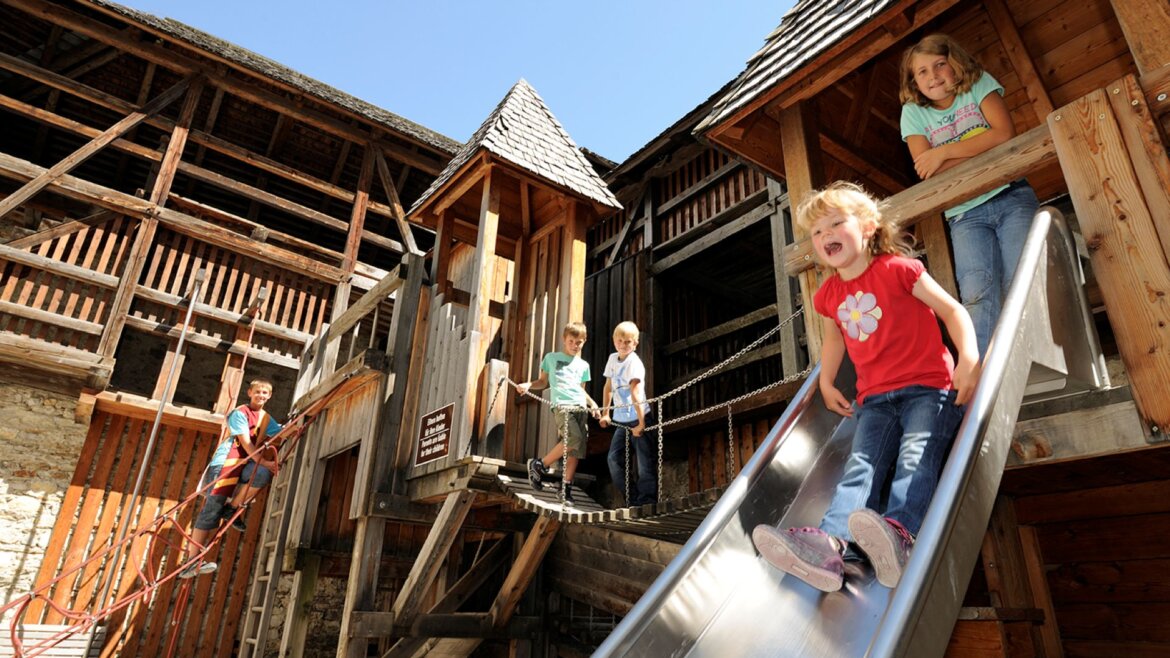 Čtyři děti si za slunečného dne hrají na dřevěném hřišti u hradu Mauterndorfu. (Zvětšený pohled)