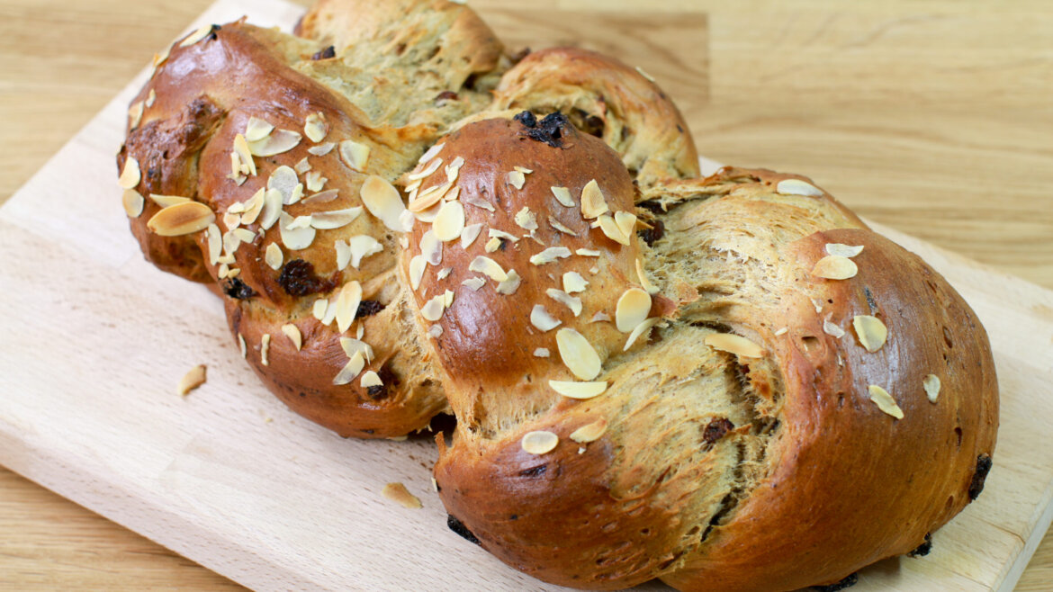 Pletený bochník chleba s rozinkami a plátky mandlí položený na dřevěném prkénku.