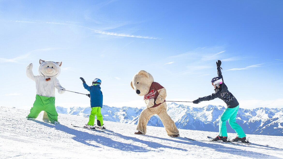 Zábava pro děti s maskotem Kogel-Mogel © Wildkogel-Arena Dva lidé ve zvířecích kostýmech a dva lyžaři se drží lana během závodu Soutěž Wildkogel-Arena Salzburgerland. (Zvětšený pohled)