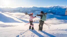 wildkogelarena_2024-5294.1920×0 Dva lyžaři s výstrojí stojí na zasněžené hoře a plácají si s malebnými vrcholky a mraky v pozadí.