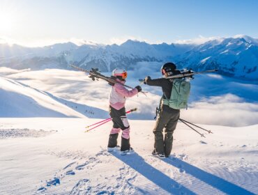 Dva lyžaři s výstrojí stojí na zasněžené hoře a plácají si s malebnými vrcholky a mraky v pozadí.