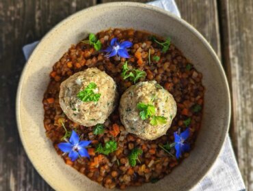 Mísa čočkového guláše se dvěma knedlíky, modrými jedlými květy a čerstvými bylinkami na dřevěném stole.