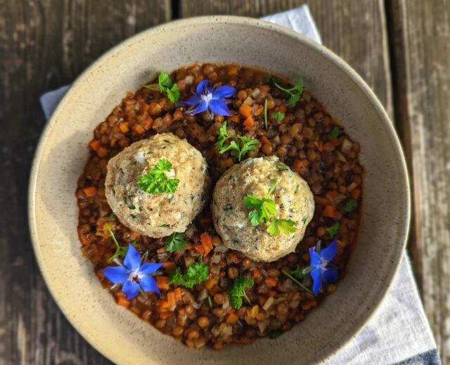 (c) Salzburger Agrar MarketingMartina Höfelmaier (2) Mísa čočkového guláše se dvěma knedlíky, modrými jedlými květy a čerstvými bylinkami na dřevěném stole.