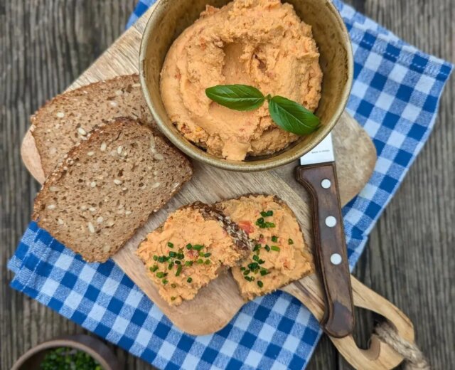 (c) Salzburger Agrar MarketingMartina Höfelmaier 3 Miska s pomazánkou s bazalkou, plátky žitného chleba a nůž na prkénku přes modrou kostkovanou látku.