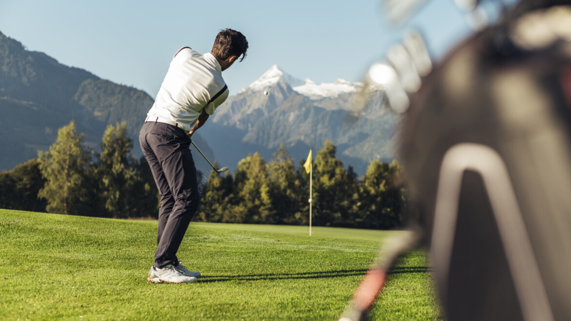 Golfista se za slunečného dne houpe na zeleném hřišti s horami a vlajkou v pozadí.