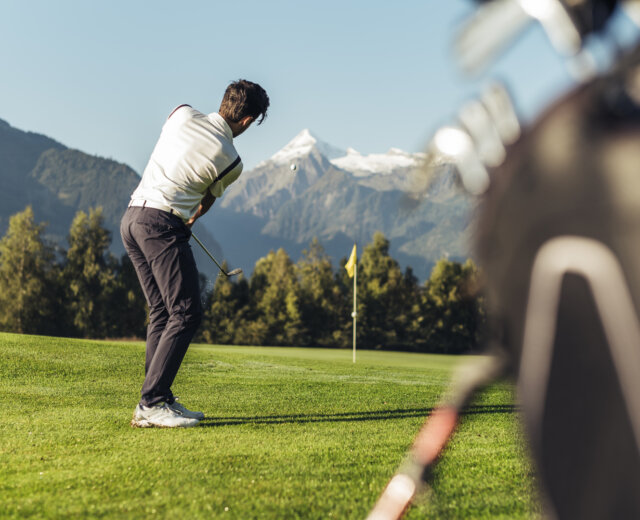 Golfista se za slunečného dne houpe na zeleném hřišti s horami a vlajkou v pozadí.
