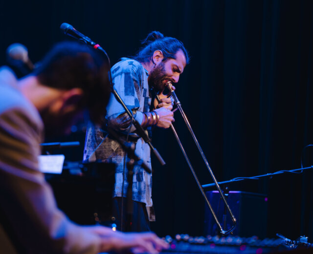 Hudebník na pódiu hraje na trombón, zatímco jiný hraje na klávesy v popředí.