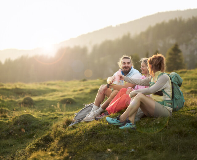 Tři lidé sedí na travnatém kopci, usmívají se a povídají si, s batohy na zádech a horami v pozadí.