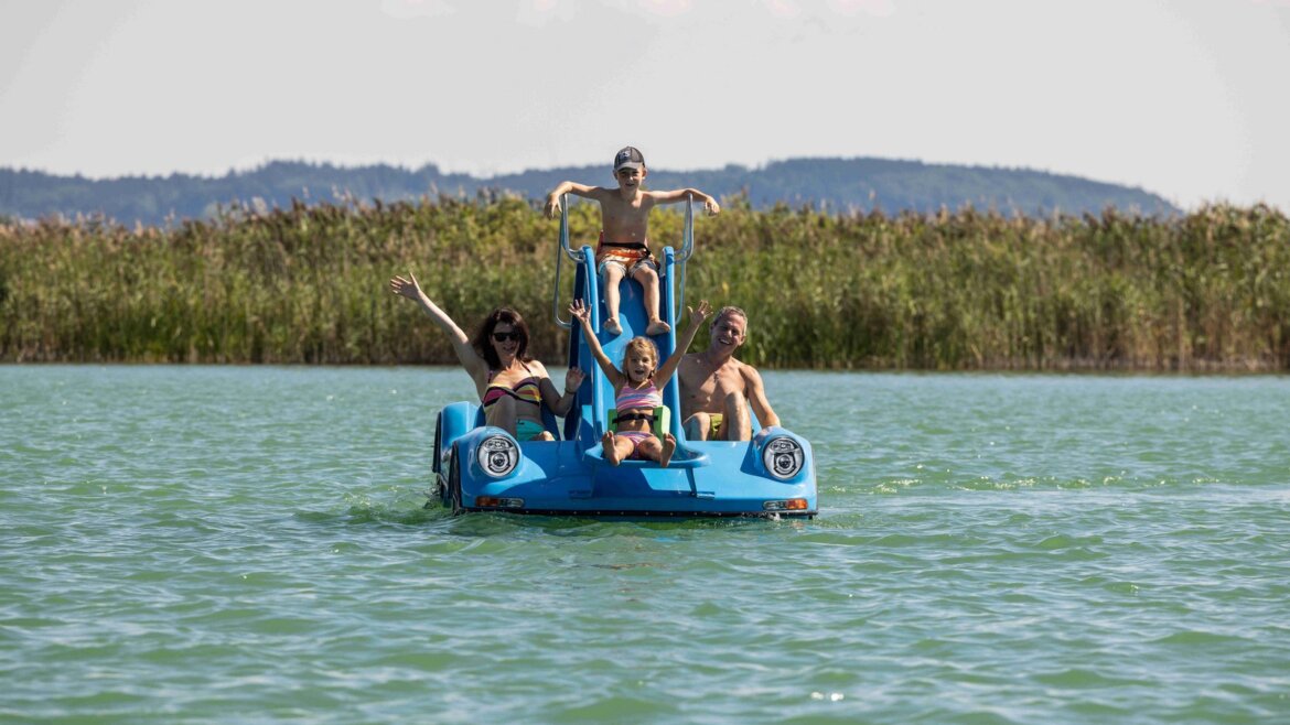 Čtyři lidé si užívají slunečný den na modrém šlapadle v oblasti Salzburger Seenland obklopeném klidnou vodou.