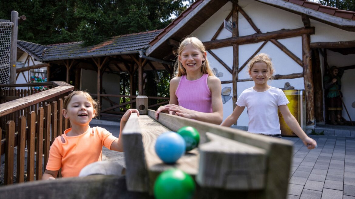 Tři děti se usmívají a hrají si s kutálejícími se míči v Salcburské zemi jezer před venkovským stavením.