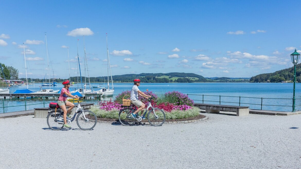 Dva lidé jedou na kolech u Salcburského jezera, kde jsou za slunečného dne vidět plachetnice, květiny a kopce.