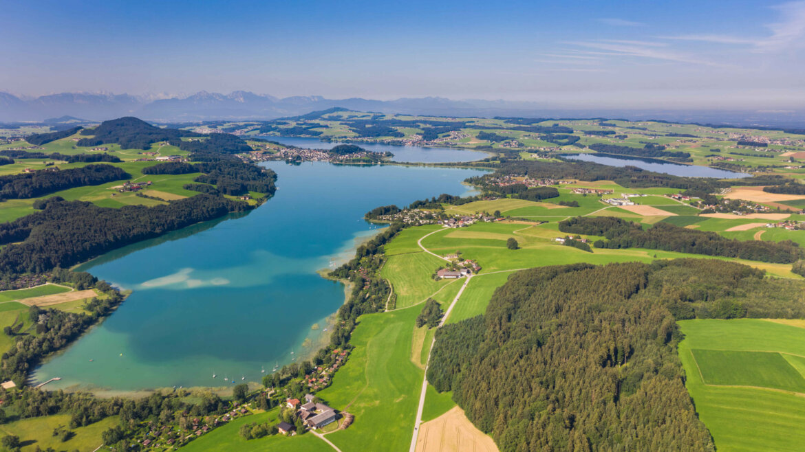 Letecký pohled na modré jezero obklopené zelenými poli, lesy a vzdálenými horami pod jasnou oblohou. (Zvětšený pohled)