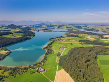 Letecký pohled na modré jezero obklopené zelenými poli, lesy a vzdálenými horami pod jasnou oblohou.