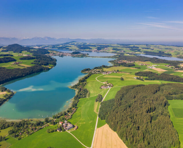 Letecký pohled na modré jezero obklopené zelenými poli, lesy a vzdálenými horami pod jasnou oblohou.