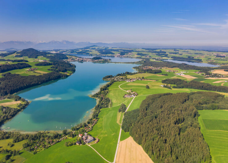 Letecký pohled na modré jezero obklopené zelenými poli, lesy a vzdálenými horami pod jasnou oblohou.
