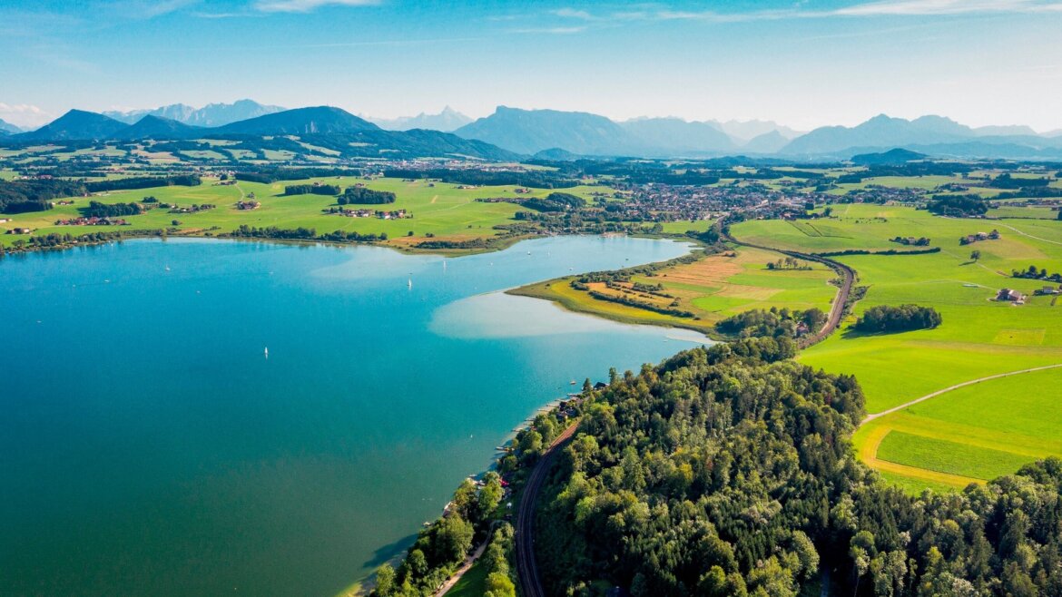 Letecký pohled na jezero Wallersee v Salcburské zemi jezer se zelenými poli, stromy a vzdálenými horami pod modrou oblohou.