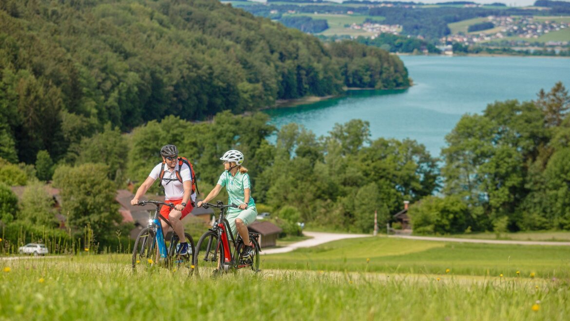 Dva lidé jedou na kolech u jezera v Salcburské zemi jezer, za nimi jsou stromy a vzdálené kopce.