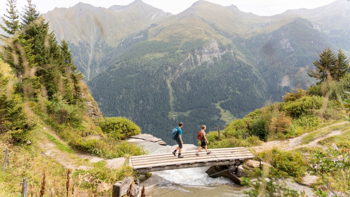 Dva turisté přecházejí dřevěný most u Bruck Fusch Großglockner v létě, za nímž jsou hory a zeleň.