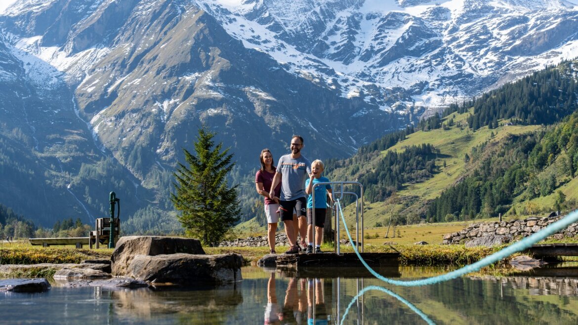 Tři lidé stojí na mostě v Bruck Fusch Großglockner v létě, za nimi jsou zasněžené vrcholky a zelené kopce. (Zvětšený pohled)