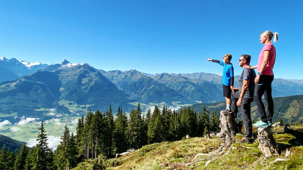 Tři lidé stojí na pařezech stromů s výhledem na malebnou horskou krajinu Piesendorf a Niedernsill v létě. (Zvětšený pohled)