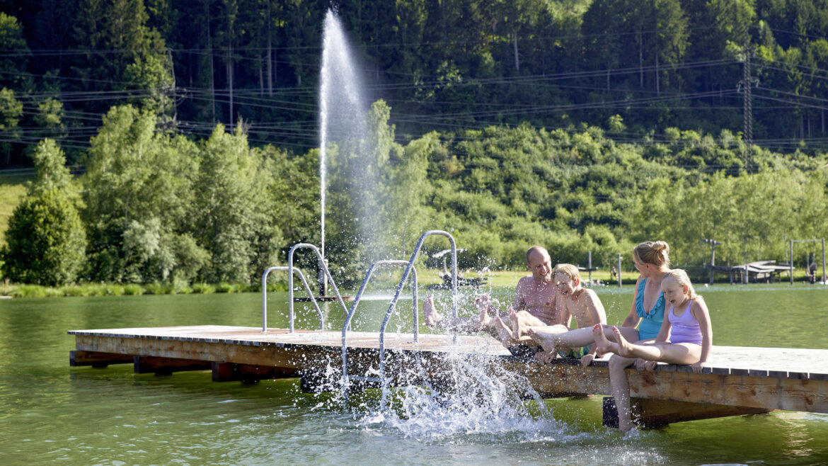 Rodina na dřevěném molu na přírodním koupališti Niedernsill s travnatými plochami a horami kolem (Zvětšený pohled)