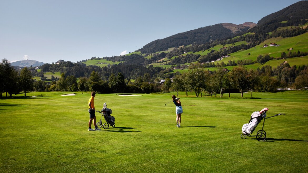 Dva lidé hrající golf na zeleném hřišti v Mittersillu v létě, v pozadí hory. (Zvětšený pohled)