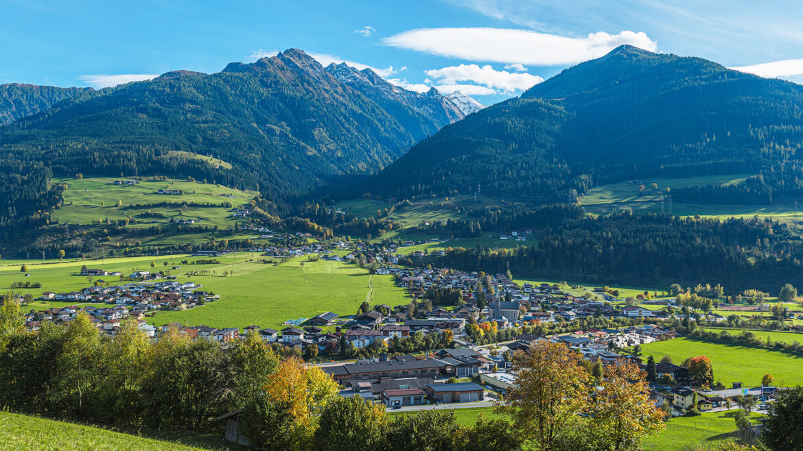 Obec Niedernsill v létě, se zelenými poli a domy v horském údolí. (Zvětšený pohled)