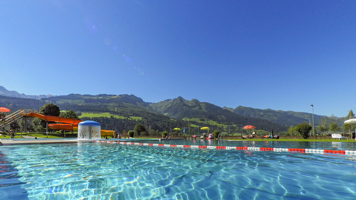 Bazén v Hinkelsteinbadu v Piesendorfu s klidnou vodní hladinou, tobogánem a horskou scenérií v pozadí (Zvětšený pohled)