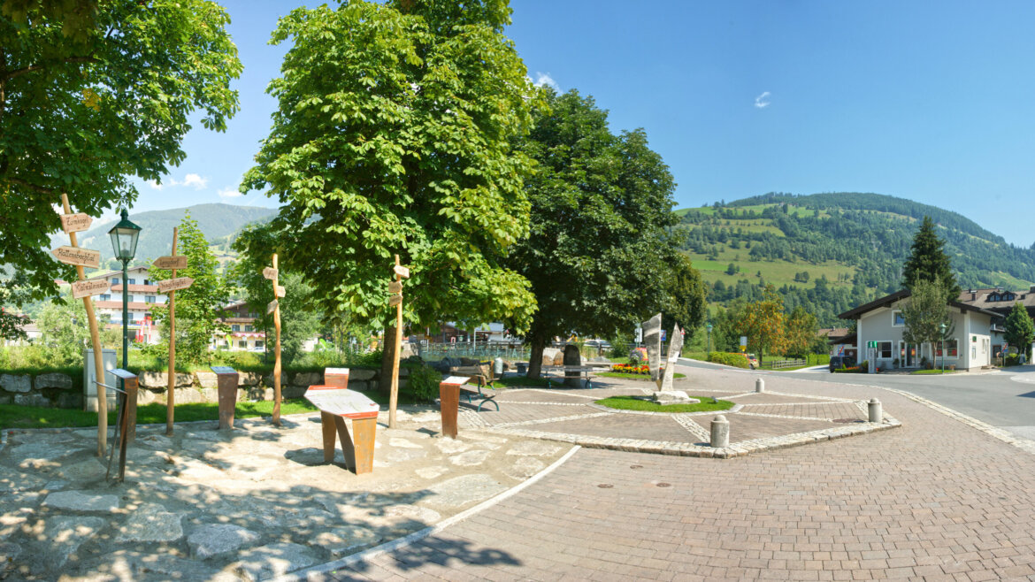 Slunné náměstí v Piesendorfu a Niedernsillu v létě se stromy, lavičkami a zelenými kopci pod modrou oblohou. (Zvětšený pohled)