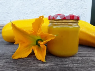 Sklenice se žlutým cuketovým džemem s víčkem s červeným kostkovaným vzorem, vedle ní leží cuketa a žlutý cuketový květ.