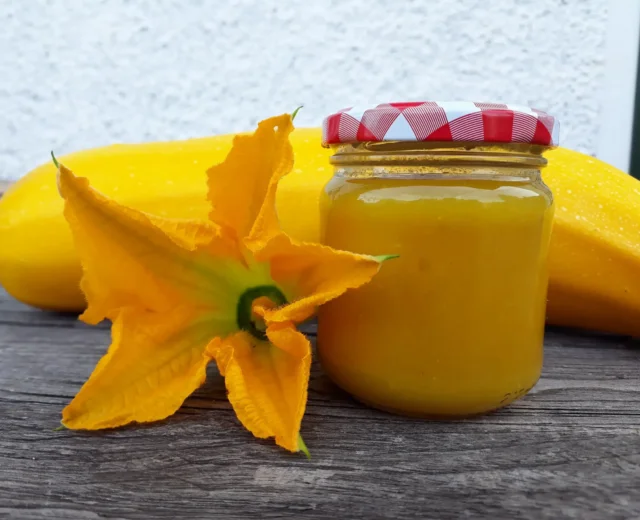 Sklenice se žlutým cuketovým džemem s víčkem s červeným kostkovaným vzorem, vedle ní leží cuketa a žlutý cuketový květ.