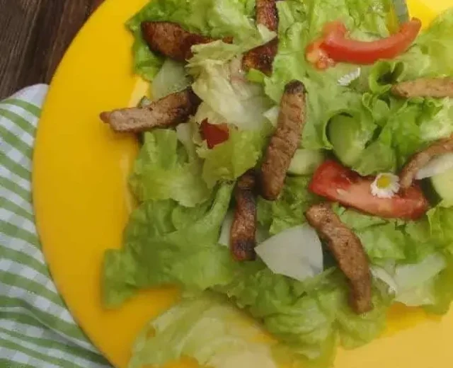 Bunter-Salat-mit-gebratenen-Hühnerbruststreifen_©SAM.jpg Na dřevěném stole stojí žlutý talíř se zeleným salátem, plátky rajčat, grilovanými proužky masa a květinami.