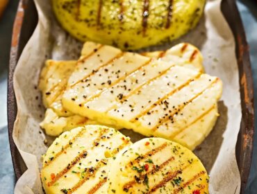 Gegrillter Käse mit Kräutermarinade © Canva.png Grilované plátky sýra halloumi a kolečka bylinkového sýra na plechu vyloženém pergamenovým papírem.