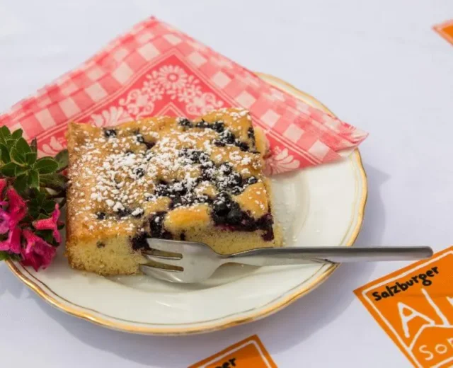 meisis-reise-lungau-schwarzbeerkuchen-auf-der-twenger-alm-1170×658.jpg Kousek bobulového dortu, klasika z letních receptů ze Salcburska, posypaný cukrem a jedlými květinami na červeném ubrousku.