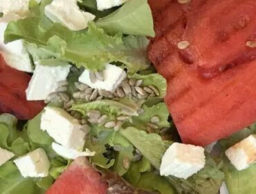 Salat-mit-gegrillter-Wassermelone_Ulli Hammerl Salát, plátky grilovaného vodního melounu, kostkami sýra a slunečnicovými semínky na bílém talíři.