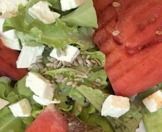 Salat-mit-gegrillter-Wassermelone_Ulli Hammerl Salát, plátky grilovaného vodního melounu, kostkami sýra a slunečnicovými semínky na bílém talíři.