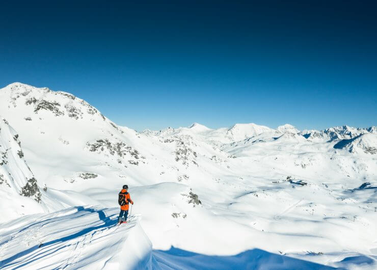 En skiløber i orange står på en snedækket piste i Obertauern med store bjergtoppe og en klar blå himmel.