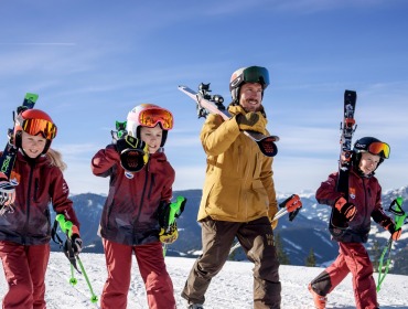 En mand og tre børn i skitøj bærer ski på et snedækket bjerg under en klar blå himmel.