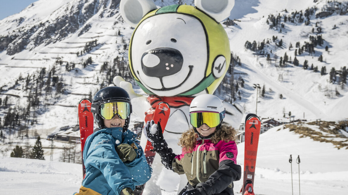 To skiløbere knæler i sneen ved en bjørnemaskotstatue, mens de nyder at stå på ski i Obertauerns sneklædte bjerge. (Forstørret visning)