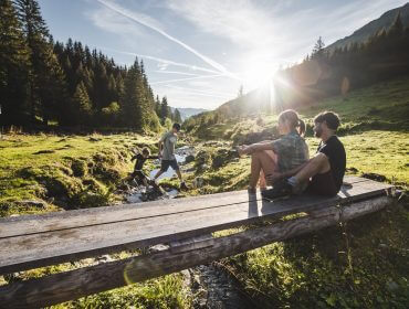 To personer sidder på en træbro i det solrige Skicircus-bjerglandskab, mens andre krydser et vandløb.