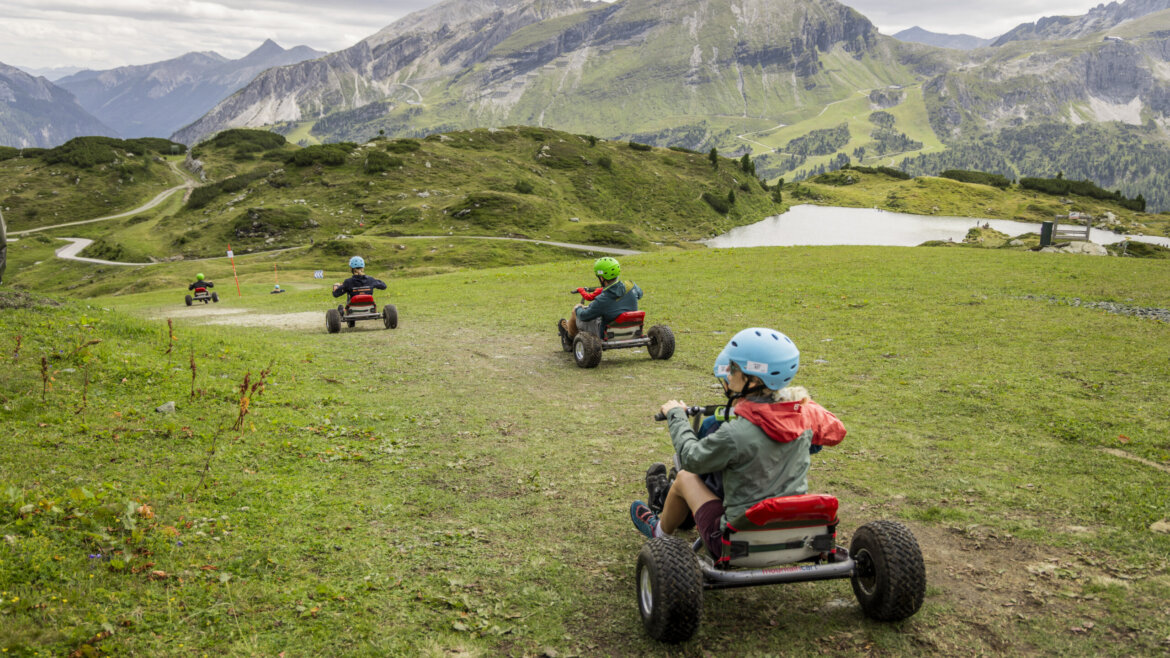 Børn kører i gokart ned ad en græsklædt bakke med bjerge og en sø - sommersjov efter skiløb i Obertauern. (Forstørret visning)